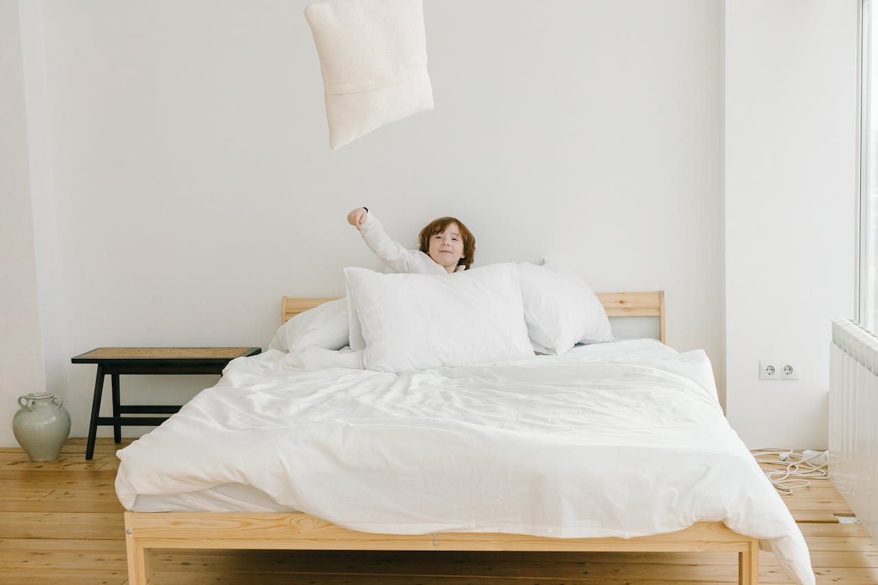 A young boy enthusiastically tosses a pillow in a bright, minimalist bedroom with white bedding.